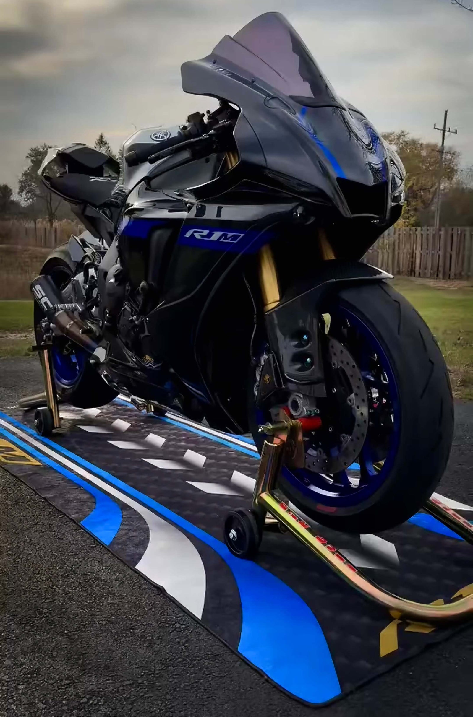 A high-performance motorcycle rests on a decorative motorcycle garage mat, featuring intricate designs in shades of blue, white, and black. The bike has gold front forks and stands on a paved outdoor surface, surrounded by a wooden fence and trees.