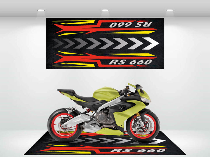 A yellow sport motorcycle with red rims is displayed on a bold Motorcycle Pit Mat for RS660, featuring vivid red, yellow, and white arrow designs and RS 660 text. Matching wall art complements the setup above the bike.