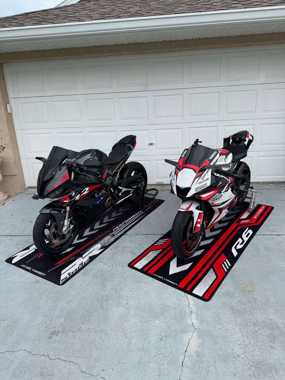 Yamaha R6 and BMW S1000RR motorcycles on custom motorcycle mats in front of a garage driveway.
