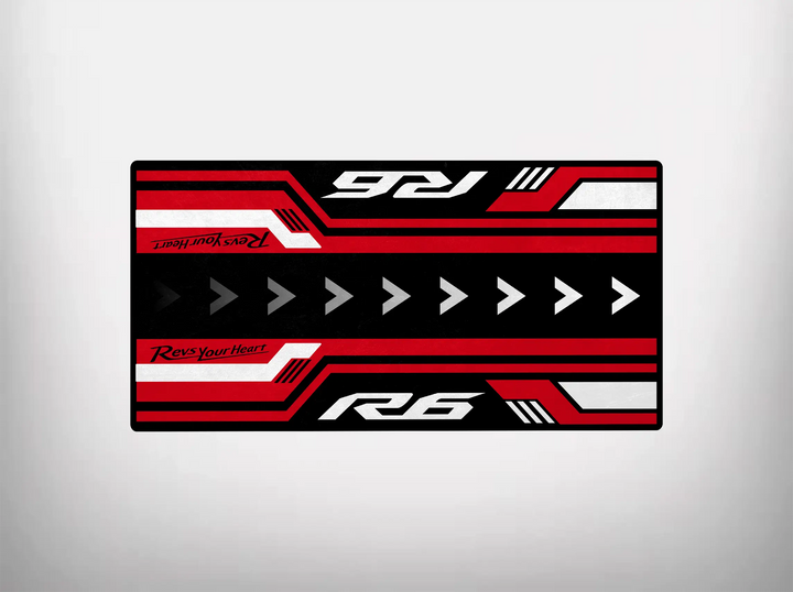 The Motorcycle Pit Mat for Yamaha R6 features a rectangular design with bold red, black, and white colors. It has right-pointing arrows with Revs Your Heart and R6 in opposite corners, offering a dynamic racing-inspired look ideal for any motorcycle garage floor.